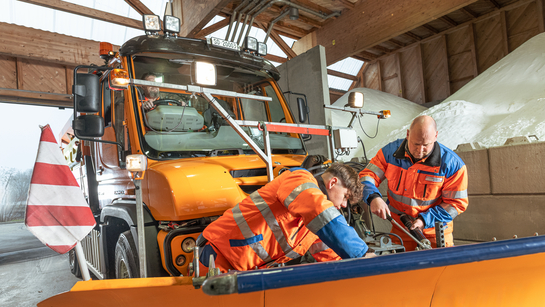 Zwei Mitarbeitende des Amts für Verkehr und Tiefbau montieren die Schneeschaufel auf dem Strassen-Räumungsfahrzeug