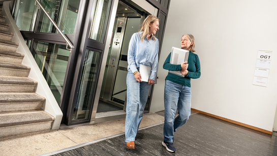Zwei Mitarbeitende des Gesundheitsamts befinden sich im Gebäude und verlassen mit dem Laptop in der Hand den Aufzug