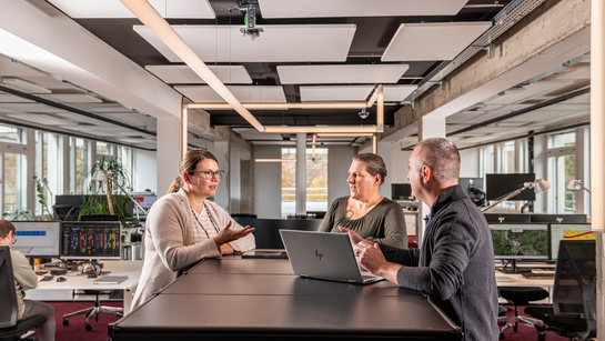 Drei Mitarbeitende beim Kurzmeeting an einem Stehpult.